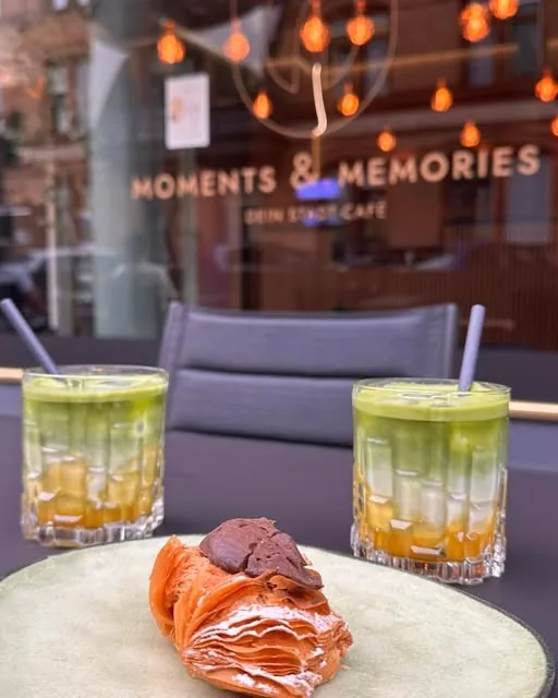 Zwei Matcha-Drinks und ein Croissant auf einem Tisch vor dem Schaufenster des Cafés