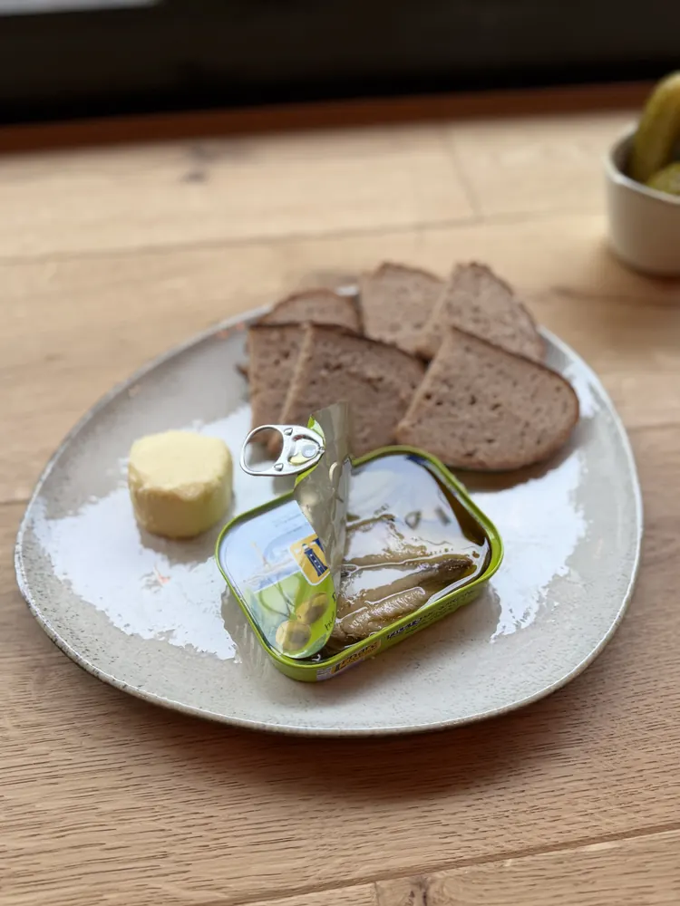 Eine geöffnete Dose Sardinen mit Butter, Brot und Gewürzgurke auf einem Teller